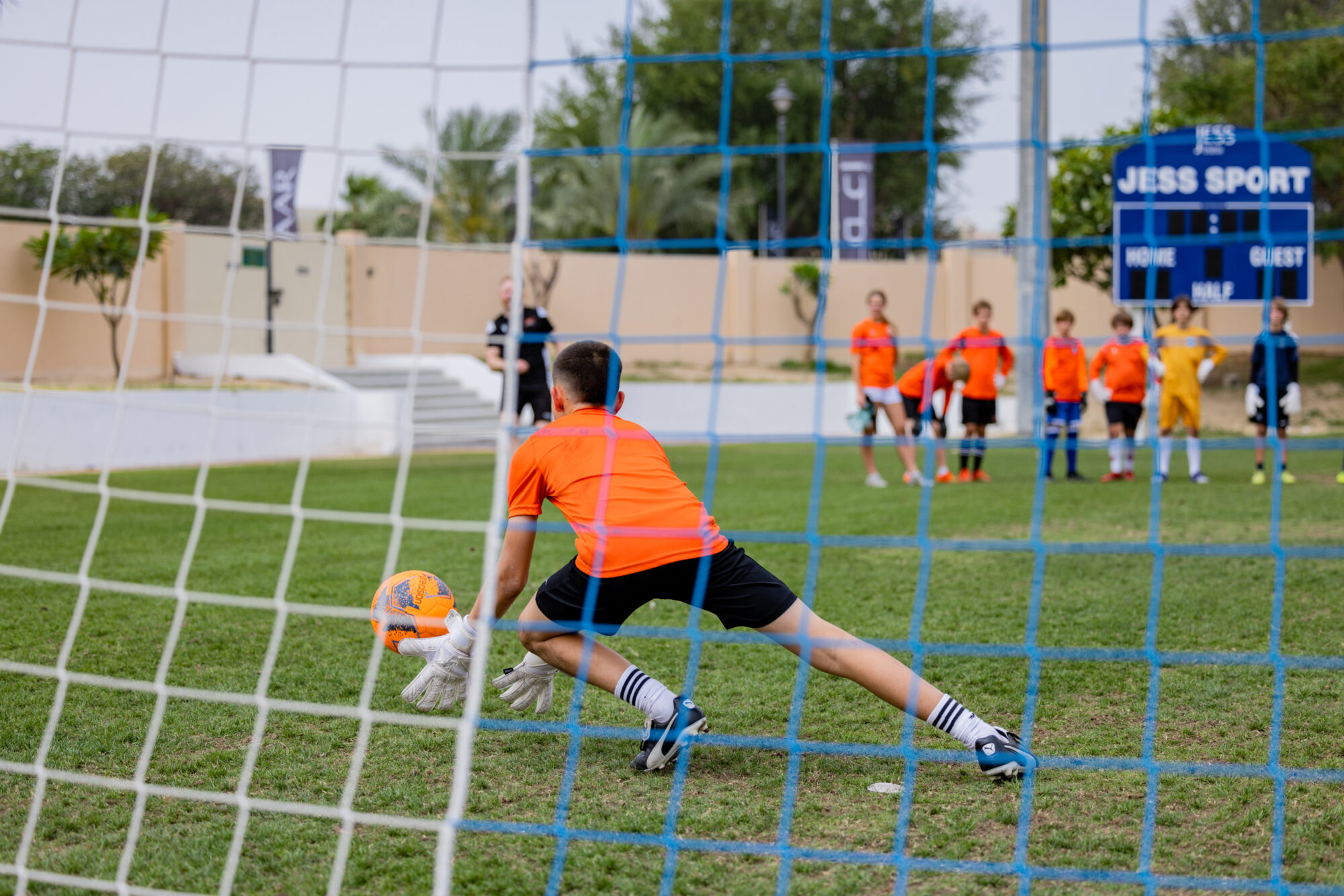 Goalkeeper in orange shirt diving to stop a ball whilst others watch